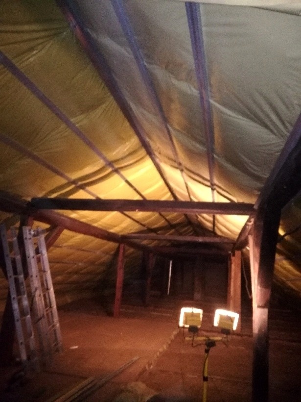 Poddasze w trakcie ocieplania, widoczne krokwie i izolacja, drabina oparta o ścianę, dwa reflektory oświetlają przestrzeń.