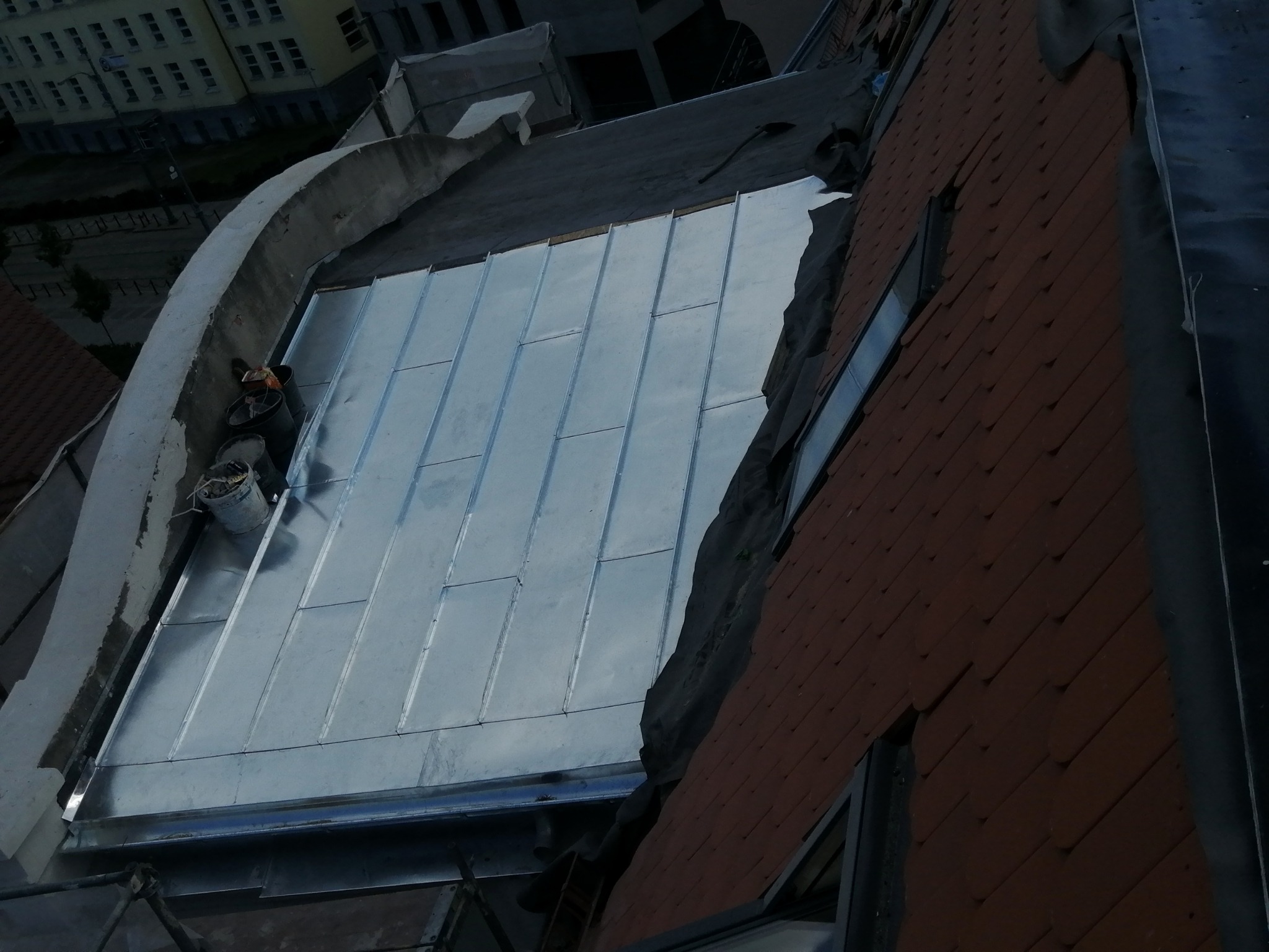 Widok z góry na fragment dachu z nowym, błyszczącym, metalowym pokryciem, obok ściana z ceglaną okładziną i okno.