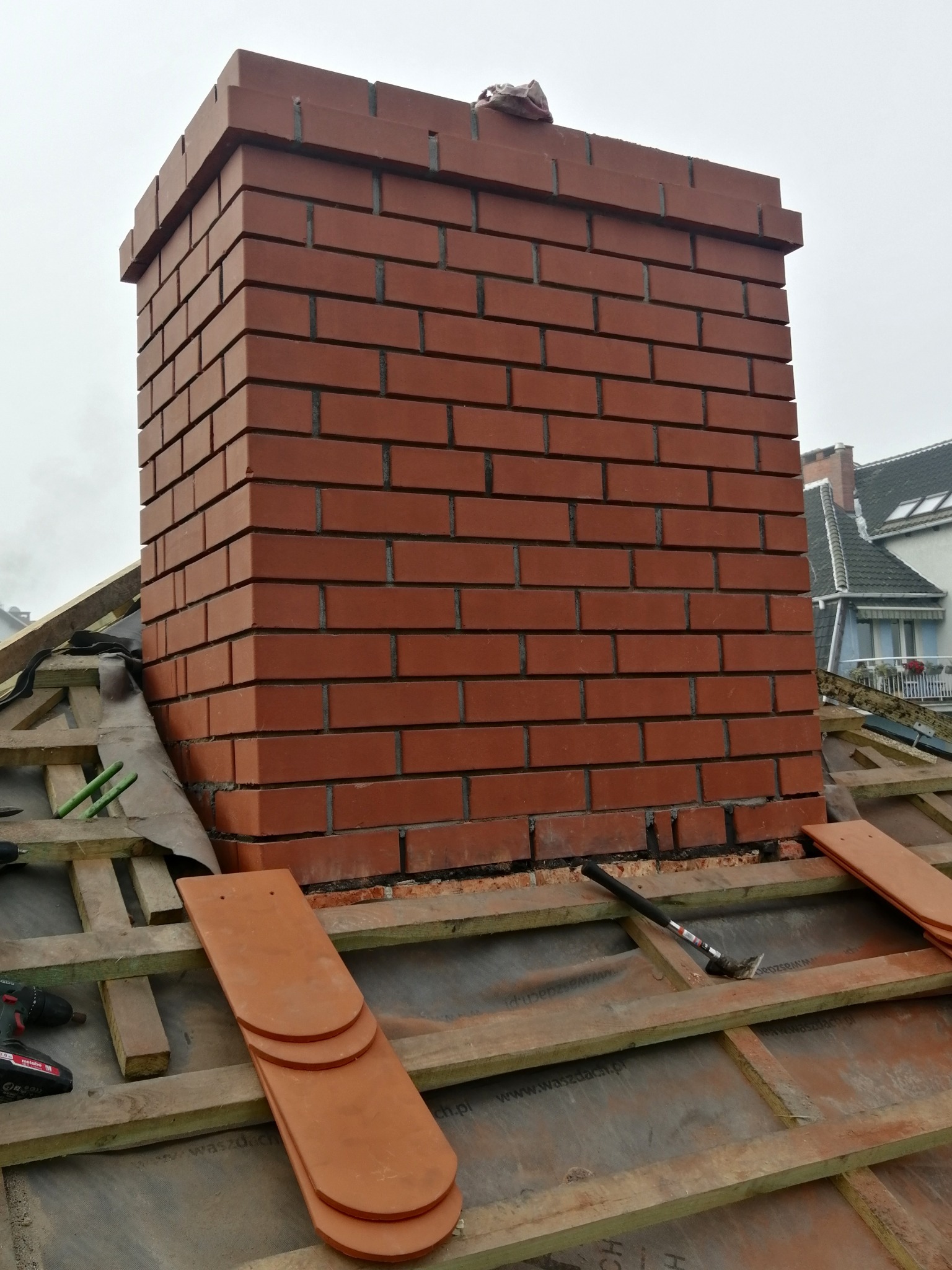 Murowany komin z czerwonej cegły na dachu w trakcie budowy, widoczne narzędzia i dachówki.