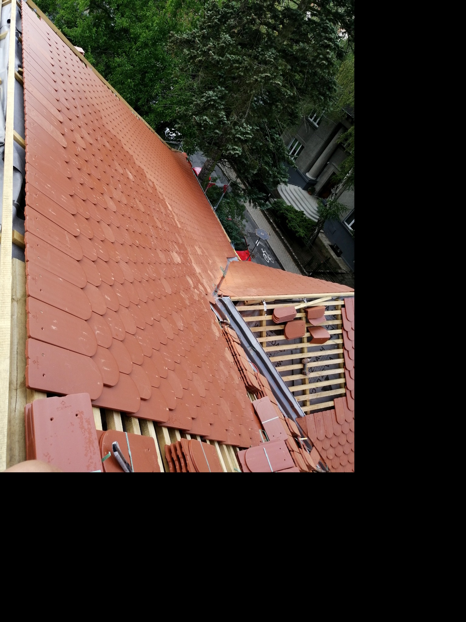 Montaż czerwonej dachówki karpiówki na drewnianej konstrukcji dachu, widoczne elementy mocujące i częściowo ukończona połać dachu, w tle zieleń drzew i budynek.