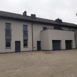 Nowoczesny szeregowiec z szarymi drzwiami wejściowymi i garażowymi, widok z zewnątrz na plac wyłożony kostką brukową, widoczne okna z balustradami i panel słoneczny na dachu.