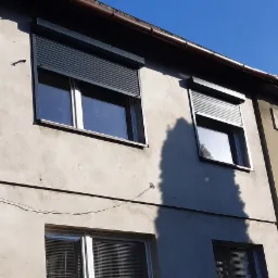 Fasada budynku z oknami zabezpieczonymi roletami zewnętrznymi, widoczny numer domu '3', cień drzewa na ścianie.