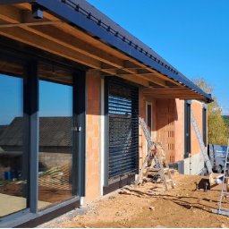 Kraft Tomasz Buczkowski - Montaż czarnych rolet zewnętrznych na oknach domu w trakcie budowy, widoczne rusztowania i ceglana elewacja. Słoneczny dzień, błękitne niebo.