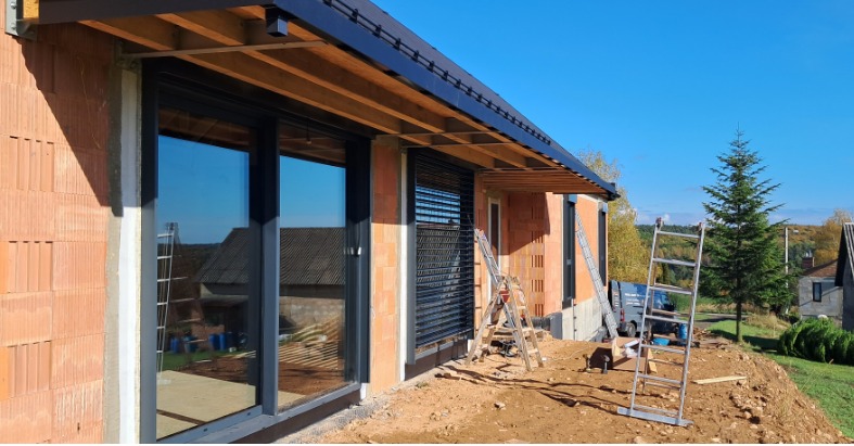 Montaż czarnych rolet zewnętrznych na oknach domu w trakcie budowy, widoczne rusztowania i ceglana elewacja. Słoneczny dzień, błękitne niebo.