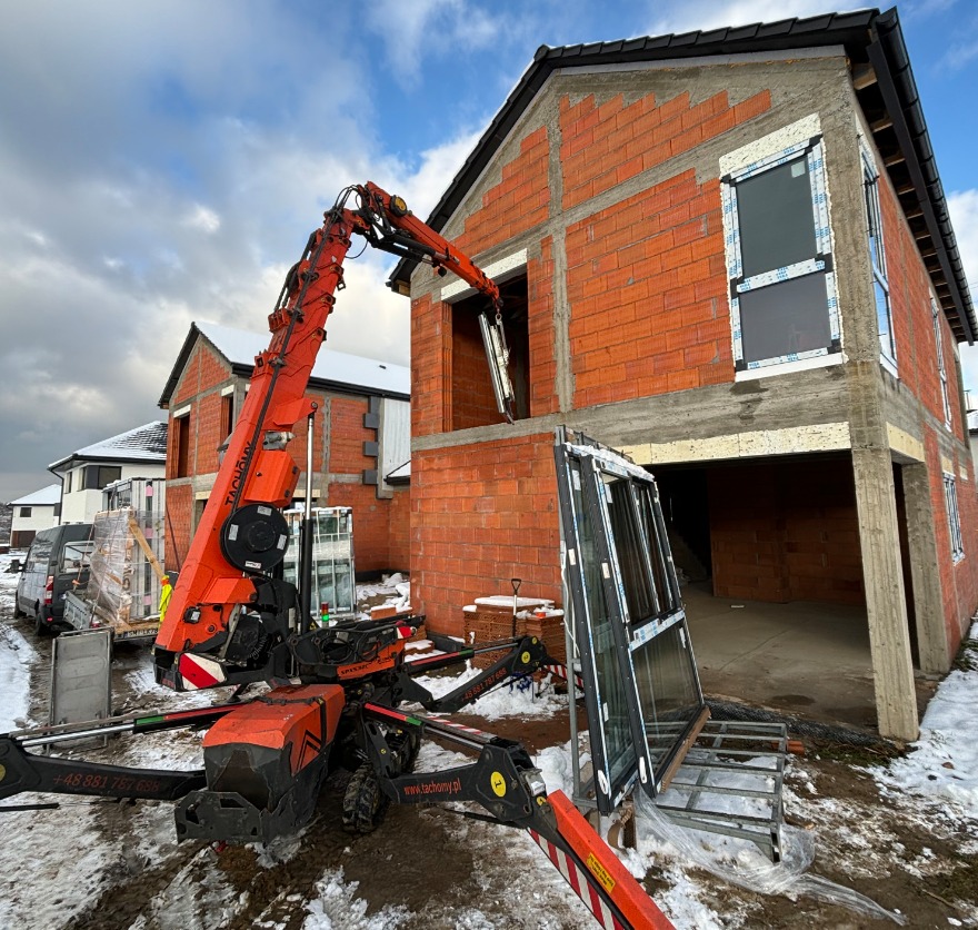 Montaż okien na budowie domu jednorodzinnego przy użyciu dźwigu. Widoczne okna na zewnątrz budynku i ceglana konstrukcja w stanie surowym.