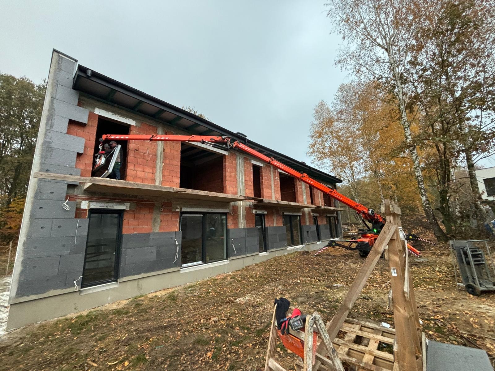 Budowa domu z czerwonej cegły, ocieplanie szarym styropianem. Widoczne czarne ramy okienne i dźwig z wysięgnikiem. Prace budowlane w toku, jesienna sceneria.