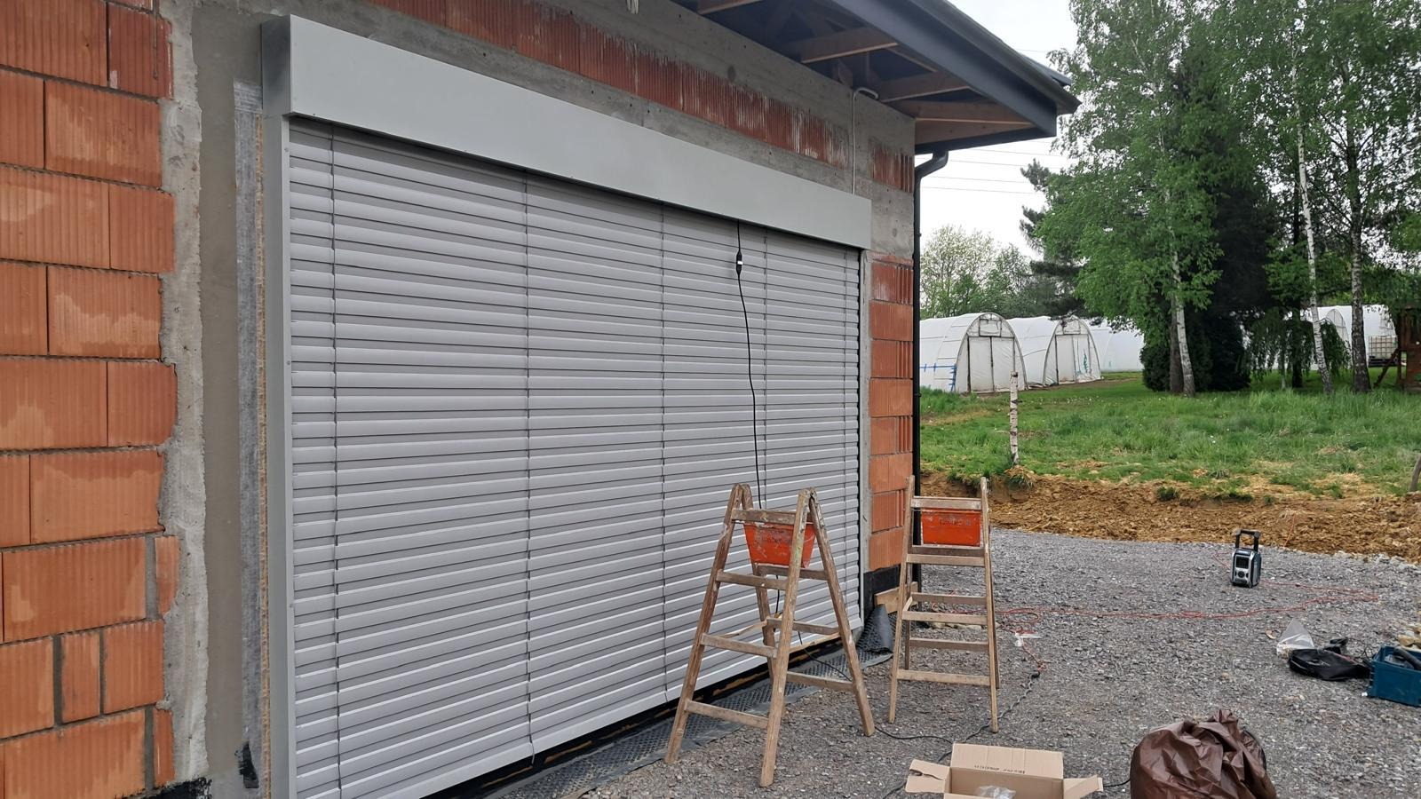 Zamknięte, szare żaluzje fasadowe na tle surowej ściany z cegły i drabiny. Widoczne szklarnie w tle. Ujęcie z dołu, podkreślające wielkość żaluzji.