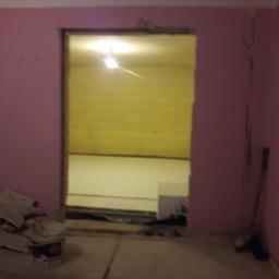 Adrian Maśny - Otwór drzwiowy w różowej ścianie, widoczny surowy stan podłogi i ściany w pokoju obok, wiadro po farbie i narzędzia w rogu.