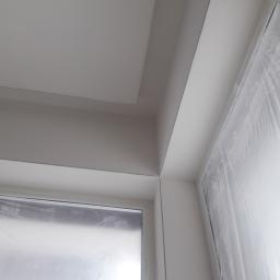 Adrian Maśny - Kąt sufitu i okna zabezpieczonego folią malarską po malowaniu, widoczne świeże, białe wykończenie.