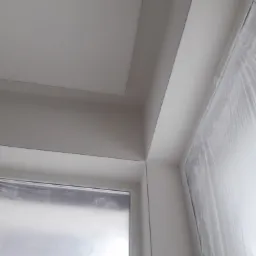 Kąt sufitu i okna zabezpieczonego folią malarską po malowaniu, widoczne świeże, białe wykończenie.