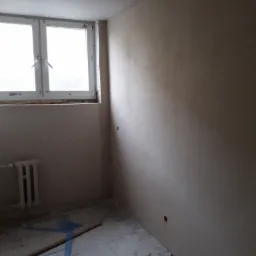 Pokój w trakcie remontu: ściany po szpachlowaniu, zabezpieczona podłoga, grzejnik żeliwny, okno z widokiem na drzewa.