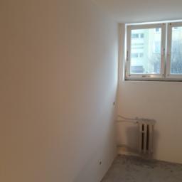 Adrian Maśny - Świeżo pomalowane na biało wnętrze pokoju z grzejnikiem pod oknem; widoczne ślady niedokończonych prac i wiadro z farbą.