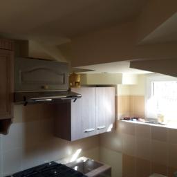 Adrian Maśny - Fragment kuchni z zabudową meblową w odcieniach beżu i szarości, widoczna płyta gazowa, okap, szafki wiszące i fragment okna z narzędziami na parapecie.