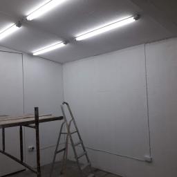 Adrian Maśny - Białe, świeżo pomalowane pomieszczenie w Puławach z widocznymi instalacjami elektrycznymi, drabina i rusztowanie malarskie w tle, gotowe do dalszych prac wykończeniowych.