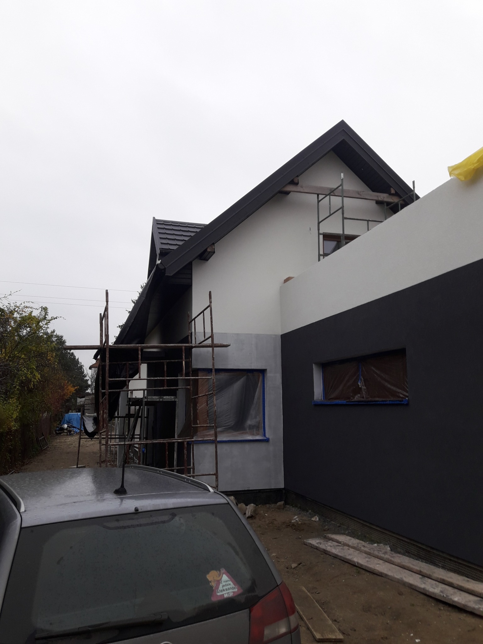 Dom jednorodzinny w trakcie malowania elewacji na biało i grafitowo, okna zabezpieczone folią, widoczne rusztowania i zaparkowany samochód z naklejką 'Z nami jedzie Aleksander'.