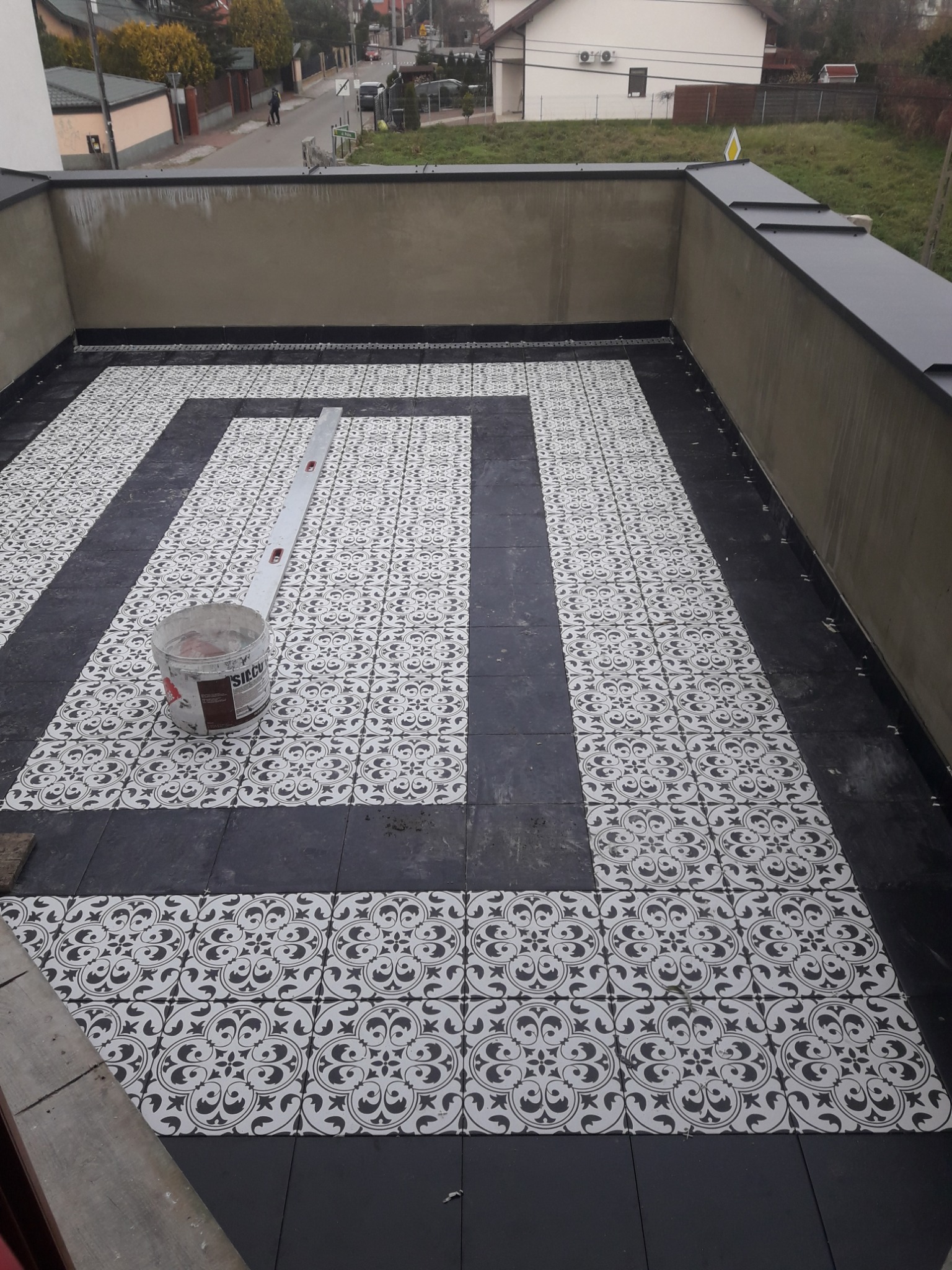 Tarasy wyłożone czarno-białymi płytkami z wzorem, widoczny kubeł z zaprawą i poziomica.