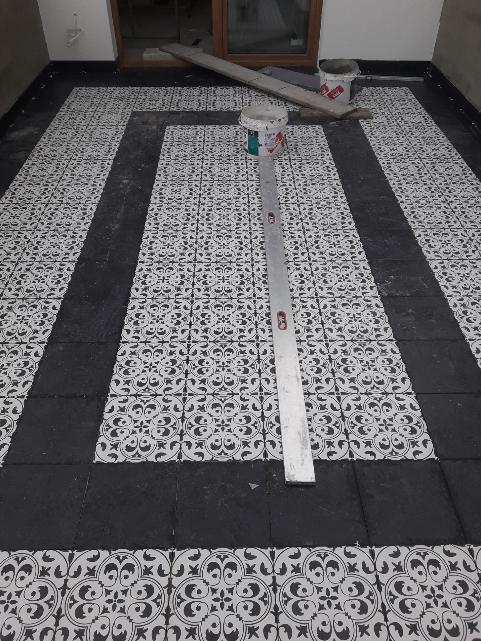 Ułożona podłoga z czarno-białych płytek o wzorzystym deseniu, z czarnymi pasami wzdłuż ścian i na środku, poziomica leży na podłodze, widoczne wiadra z klejem i deski w tle, w trakcie prac...