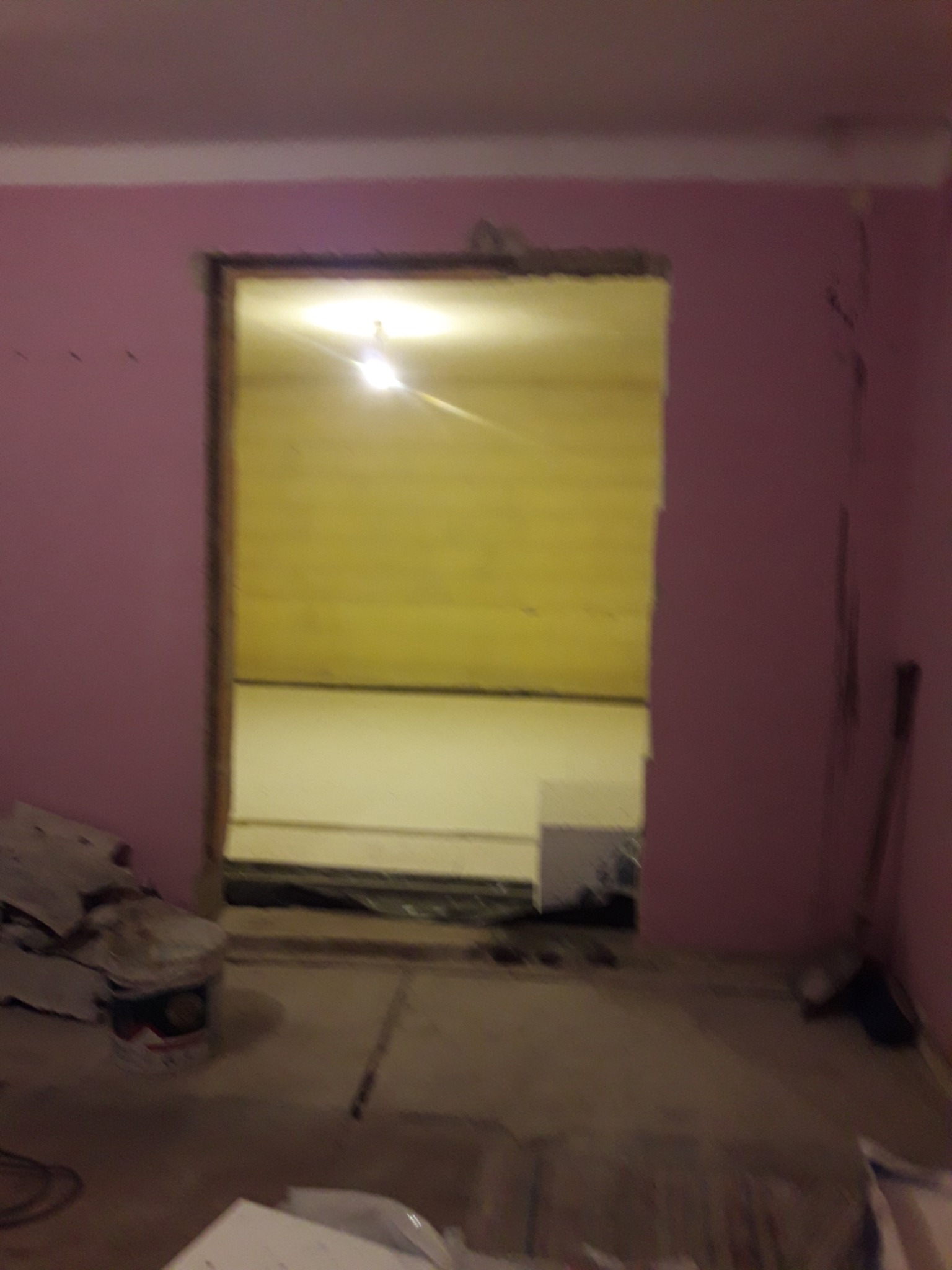 Otwór drzwiowy w różowej ścianie, widoczny surowy stan podłogi i ściany w pokoju obok, wiadro po farbie i narzędzia w rogu.