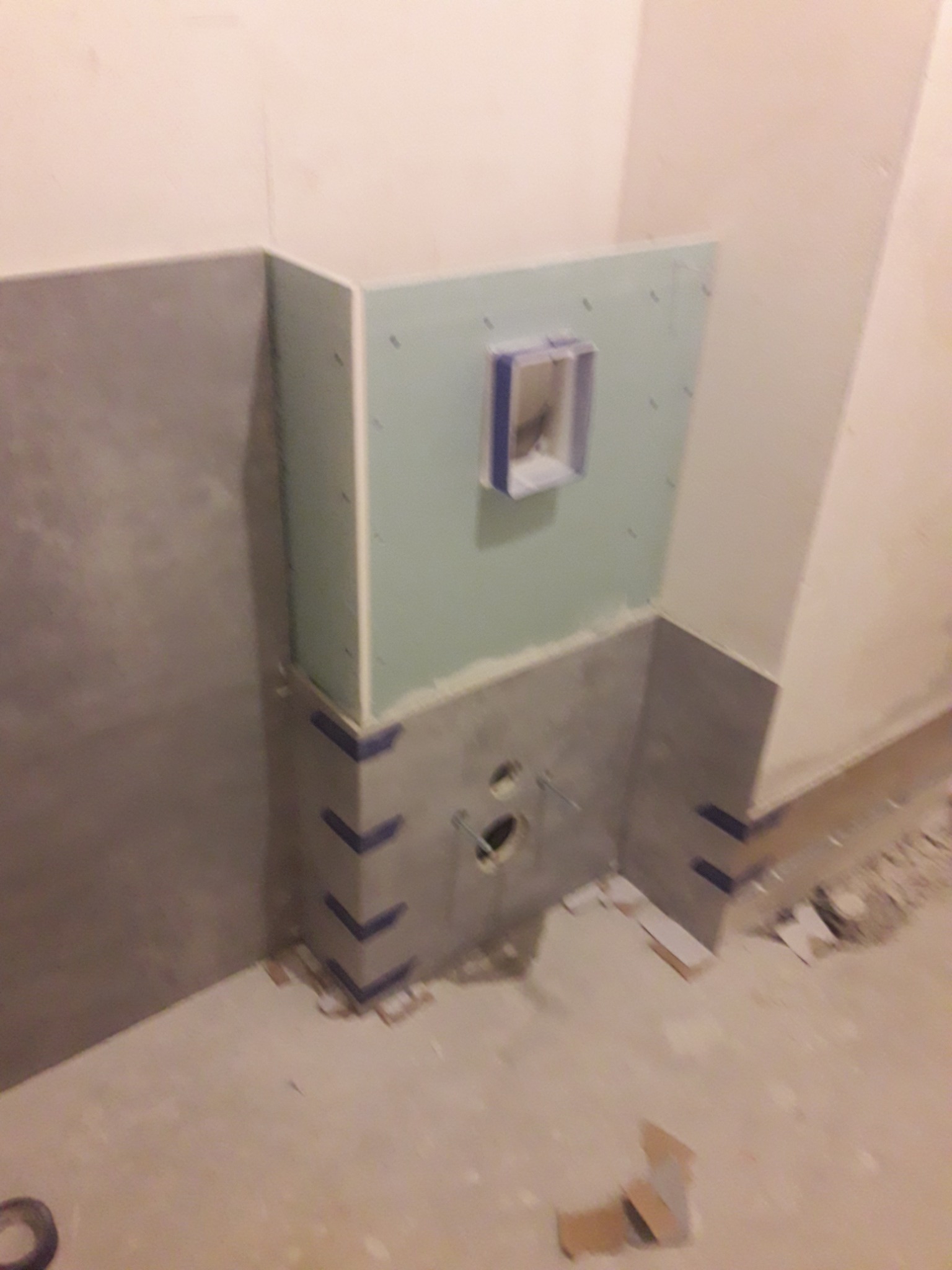 Fragment łazienki w trakcie prac wykończeniowych: częściowo ułożone szare płytki, niebieska taśma malarska zabezpieczająca krawędzie, otwór na instalację hydrauliczną i wnęka z niebieską ramką...