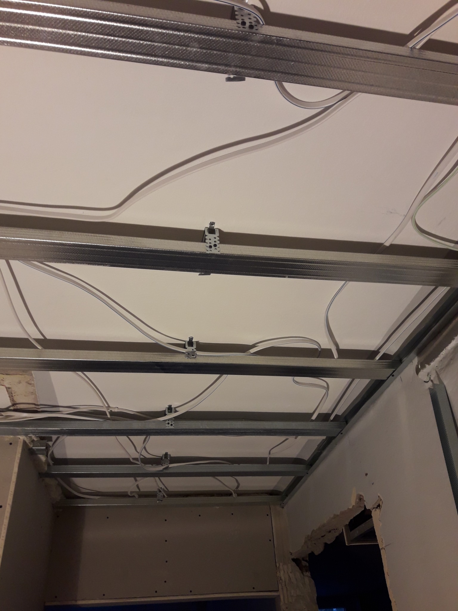 Metalowa konstrukcja sufitu podwieszanego z widocznymi kablami elektrycznymi, częściowo zamontowane płyty gipsowo-kartonowe, widok z dołu na niedokończony montaż.