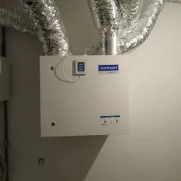 Centrala wentylacyjna Komfovent Domekt z podłączonymi aluminiowymi przewodami wentylacyjnymi, widoczny panel sterowania z wyświetlanymi danymi.