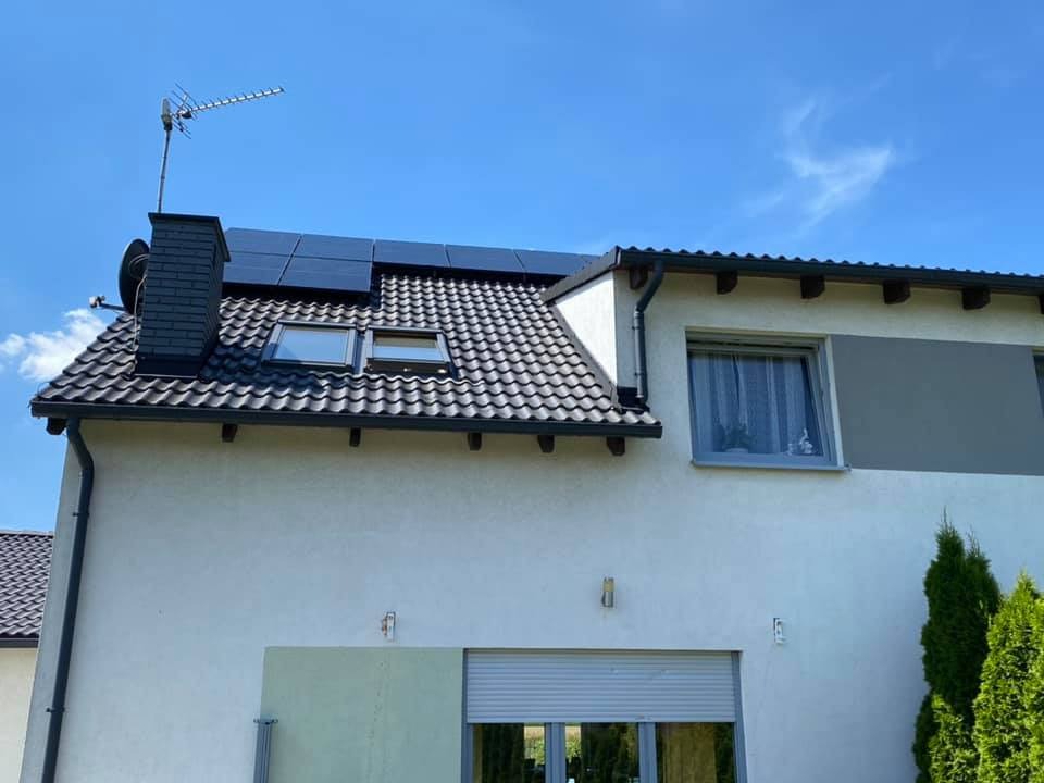 Panele słoneczne na dachu domu jednorodzinnego z czarną dachówką, kominem i anteną telewizyjną, widoczne okna dachowe i elewacja budynku w jasnym kolorze.