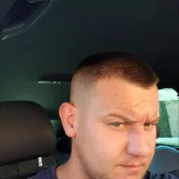 Mężczyzna z krótką fryzurą typu buzz cut siedzący w samochodzie, widoczne elementy wnętrza pojazdu.