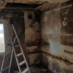 dudex - Wnętrze w trakcie remontu: odsłonięte cegły, resztki tynku na ścianach, drabina oparta o ścianę przy oknie, widoczne instalacje i uszkodzony sufit.