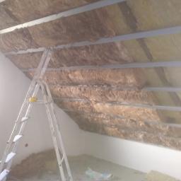 dudex - Montaż izolacji z wełny mineralnej na poddaszu, widoczne metalowe profile i drabina, resztki materiału na podłodze.