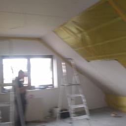 dudex - Żółta izolacja termiczna na ukośnym suficie poddasza, widoczna aluminiowa drabina i sylwetka osoby przy oknie.