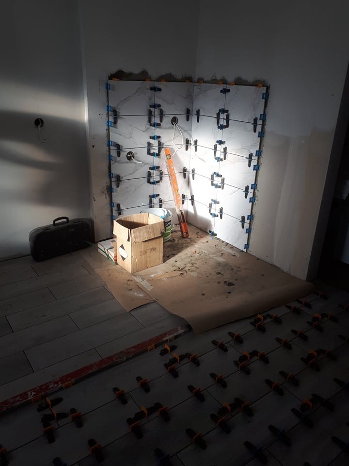 Układanie płytek marmuropodobnych na ścianie w rogu pomieszczenia, z użyciem poziomicy i systemu poziomowania płytek. Na podłodze widoczne płytki i elementy poziomujące.