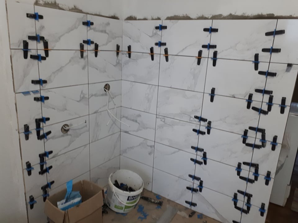 Ściana w trakcie układania płytek imitujących marmur, z widocznymi systemami poziomowania i krzyżykami dystansowymi; w tle wiadro z narzędziami.