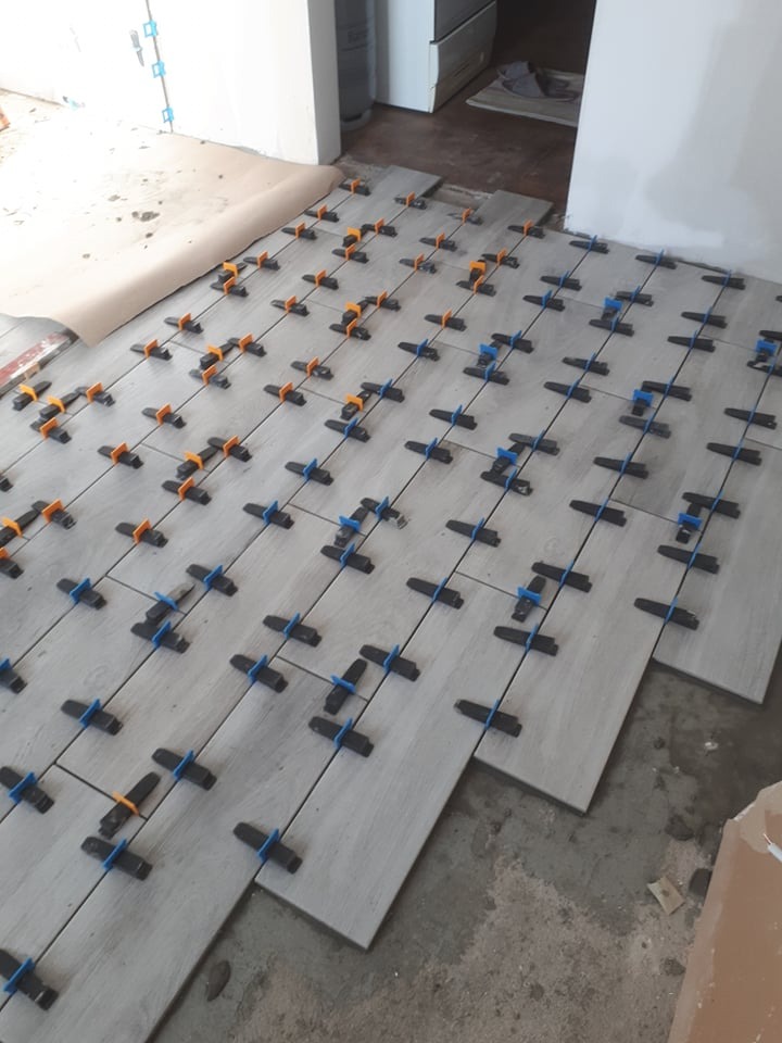 Układanie jasnoszarych płytek podłogowych z użyciem systemów poziomowania, widoczne plastikowe klipsy i kliny, surowy beton w tle.