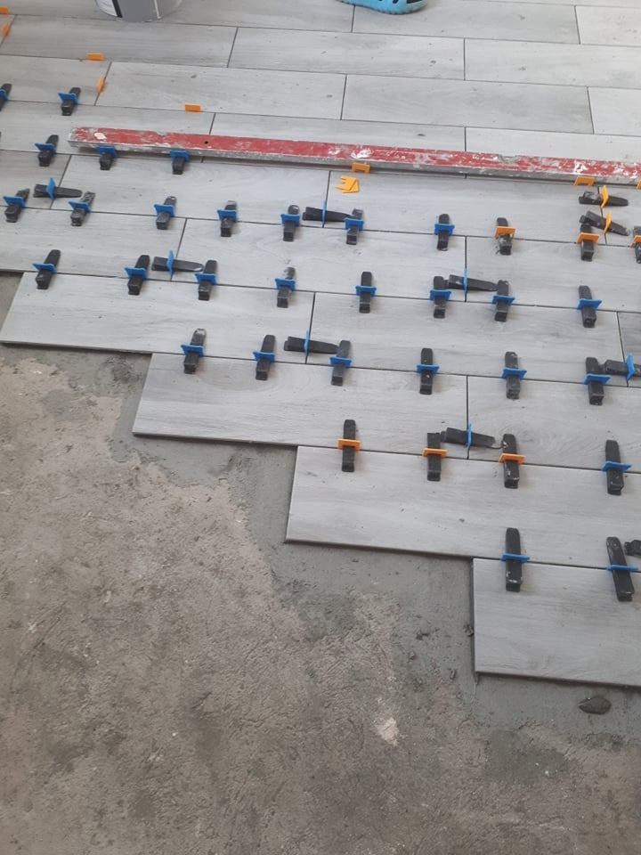 Układanie szarych płytek podłogowych z użyciem poziomicy i klipsów poziomujących, widoczny surowy beton podłoża.