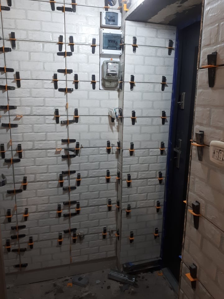 Ściana wyłożona białymi płytkami imitującymi cegłę, z użyciem systemu poziomowania płytek (klipsy i kliny), widoczne także skrzynki elektryczne i licznik.