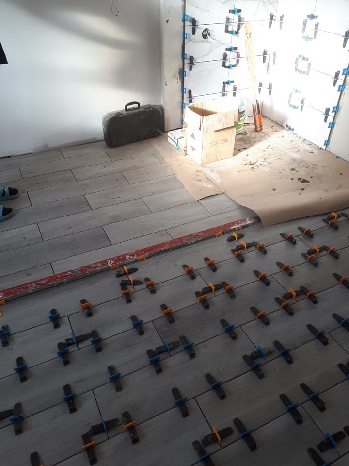 Układanie płytek podłogowych z użyciem systemu poziomowania, widoczna poziomica, na ścianie częściowo położone płytki z wzorem, w rogu stara walizka.