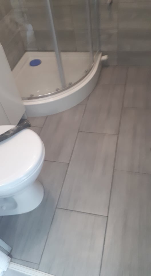 Szare płytki podłogowe w łazience z kabiną prysznicową z niebieskim odpływem i białą toaletą z szarą gumową uszczelką na górze.