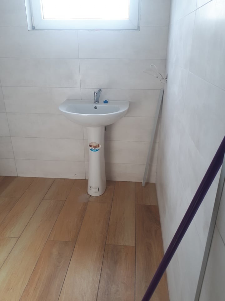 Umywalka na postumencie w łazience wyłożonej jasnymi, podłużnymi płytkami, z drewnopodobną podłogą i widocznym w rogu drążkiem do zasłony prysznicowej.