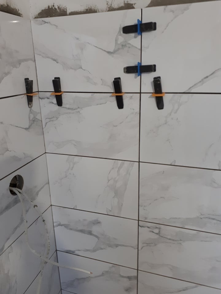 Ściana w trakcie układania płytek imitujących marmur, z widocznymi krzyżykami dystansowymi i otworem na instalację elektryczną.