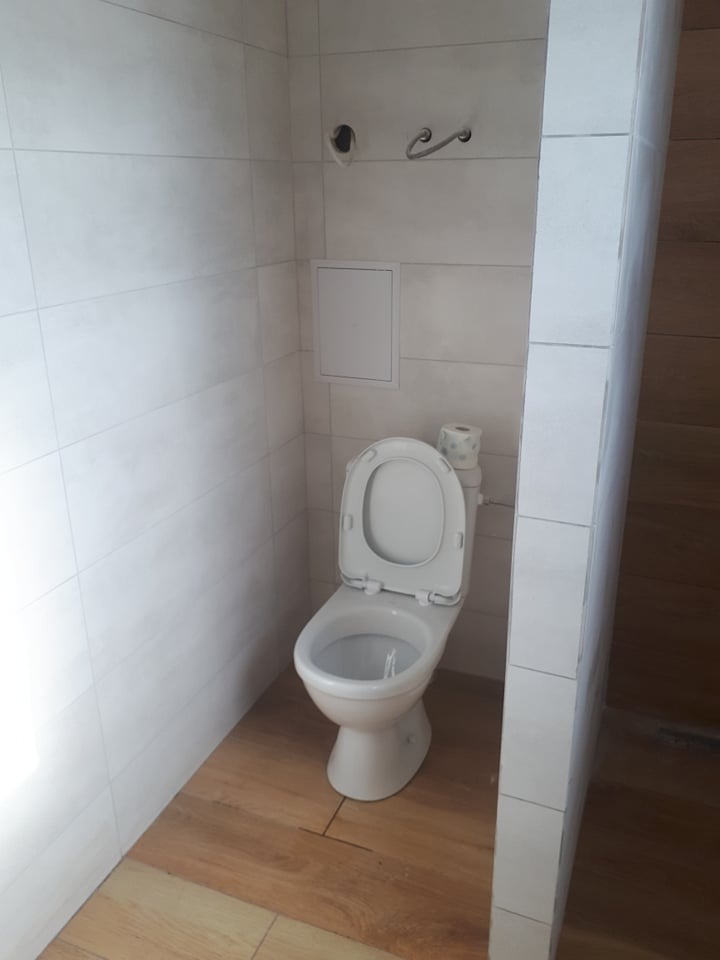 Biała toaleta z otwartą klapą na drewnopodobnej podłodze, białe kafelki na ścianach, uchwyt na papier toaletowy i rolka papieru widoczne w tle.