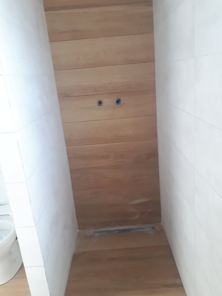 Wykończona kabina prysznicowa z drewnopodobnymi płytkami ułożonymi poziomo, widoczne dwa otwory na baterię i odpływ liniowy.