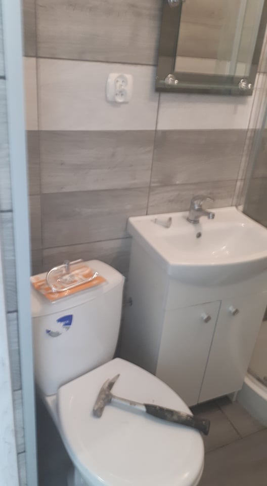 Ukończona łazienka z szarymi płytkami, umywalką z szafką, toaletą z deską sedesową, na której leży młotek.