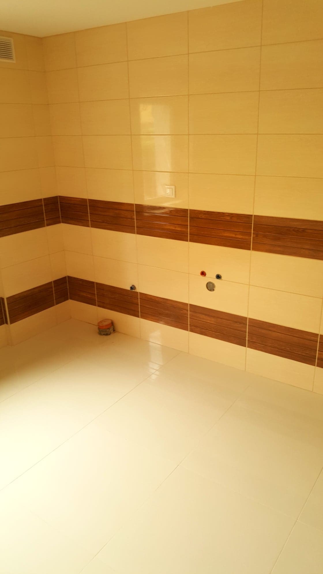 Wykończona łazienka z beżowymi płytkami i dekoracyjnym pasem z płytek imitujących drewno, widoczne przygotowane przyłącza wodne i kanalizacyjne, podłoga wyłożona jasnymi płytkami, mała puszka...