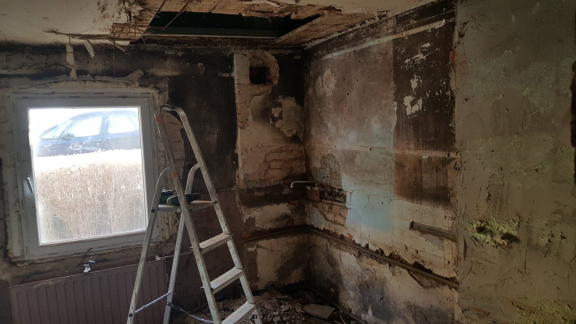 Wnętrze w trakcie remontu: odsłonięte cegły, resztki tynku na ścianach, drabina oparta o ścianę przy oknie, widoczne instalacje i uszkodzony sufit.