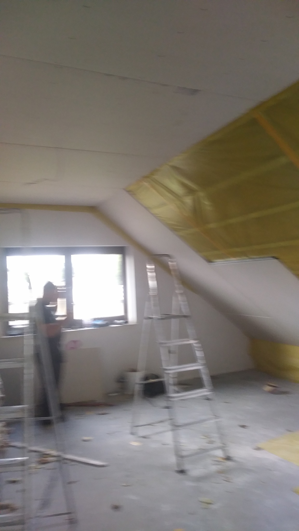 Żółta izolacja termiczna na ukośnym suficie poddasza, widoczna aluminiowa drabina i sylwetka osoby przy oknie.