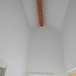 Wysoki, jasny pokój po malowaniu, z drewnianą belką stropową i oknami w dolnej części kadru.