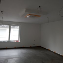 Tp bud - Pokój w trakcie remontu: szare płytki na podłodze, ściany pomalowane na biało, widoczne wystające kable, okno z żaluzjami, zabezpieczona lampa sufitowa.