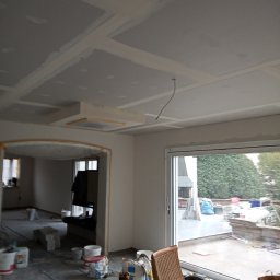 Tp bud - Wnętrze w trakcie remontu: sufit z płyt gipsowo-kartonowych ze szpachlowanymi łączeniami, folia ochronna na podłodze, worki z materiałami budowlanymi i krzesło wiklinowe w rogu pokoju.