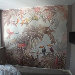 Tp bud - Ściana z fototapetą przedstawiającą tropikalny motyw z tukanami i papugami, widoczna częściowo zasłonięta folią podłoga i fragment regału na ścianie obok okna.