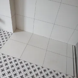 Wykończona łazienka z białymi kwadratowymi płytkami ściennymi i podłogą z dekoracyjnymi, czarno-białymi płytkami w kwiatowy wzór, widoczne łączenie płytek pod kątem.